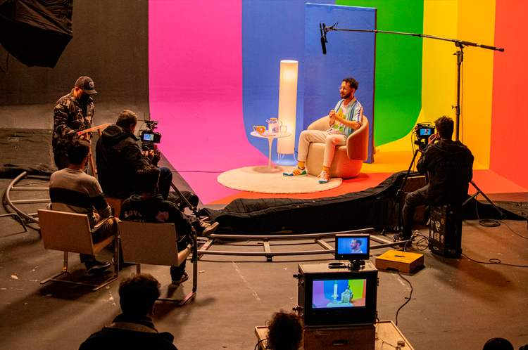 Foto do ilustrador Gutierre Sousa sendo entrevistado. Ele está sentado em uma poltrona bege clara em meio a uma pintura de arco-íris que parte do chão até o teto