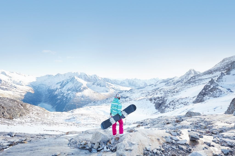 Back view of female snowboarder wearing colorful helmet, blue jacket, grey gloves and pink pants standing with snowboard in one hand and enjoying alpine mountain landscape - winter sports concept