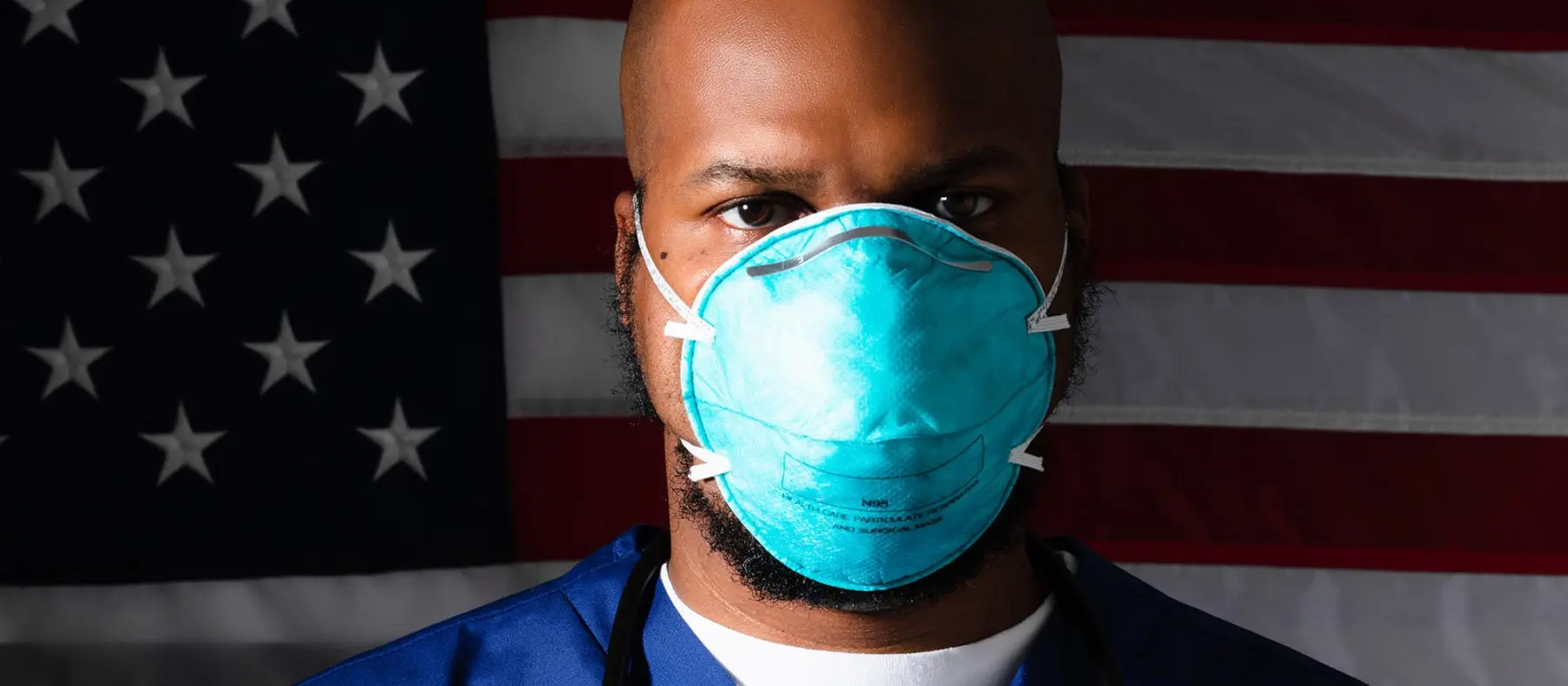 A healthcare worker wearing a medical protective mask stands in front of the american flag looking directly at the camera