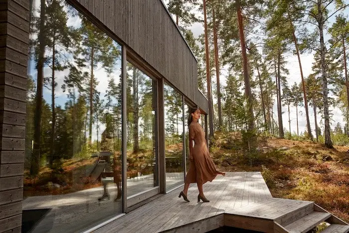 A model poses on the front porch of a wooden home built in the middle of a forest.