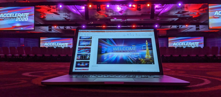 A PowerPoint slidedeck is visible on a laptop and being presented on screens at a conference venue.