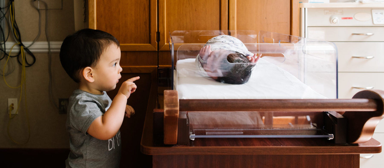 A young toddler looks in on his sibling in his hospital crib