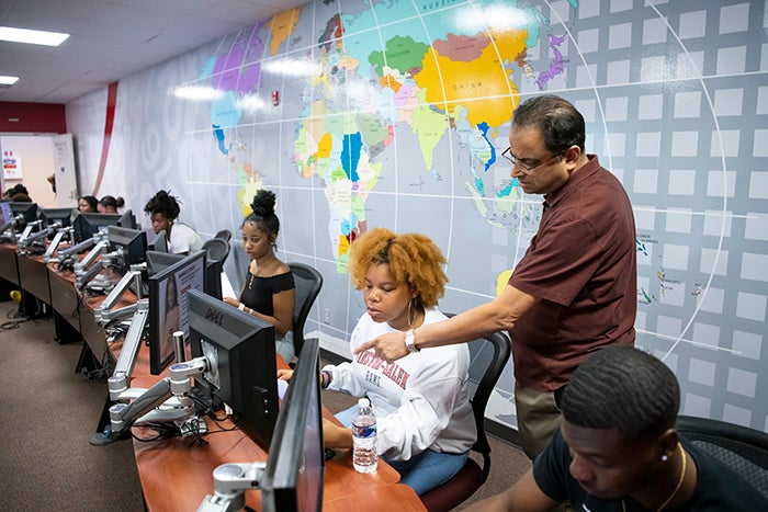 A professor stands behind a student and points to her computer screen to help her.