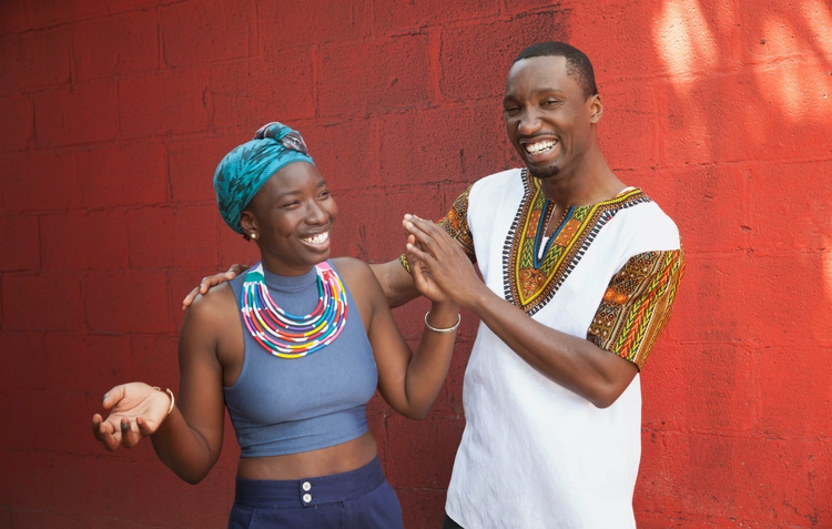 Two black people smiling by a red wall
