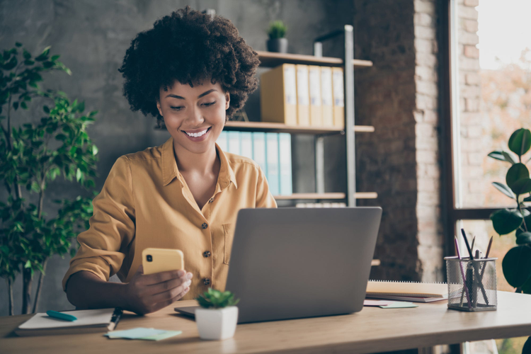 Women looking sitting at a desk while looking at her phone with her computer open. 