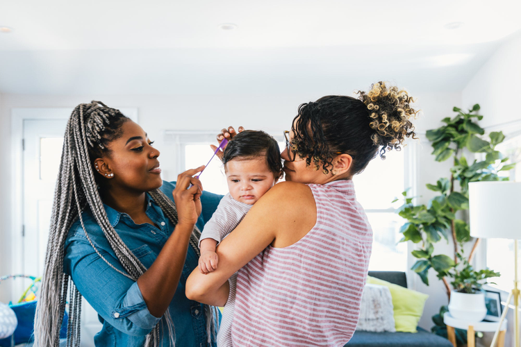 Woman holding baby with another woman combing iit's hair. 