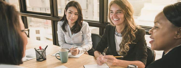 Four Women sitting at a conference table interacting.