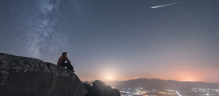 Silouhette of a man sitting on a mountain edge in the dawn, looking up at the star spangled sky, watching a shooting star above a city down in the valley below.