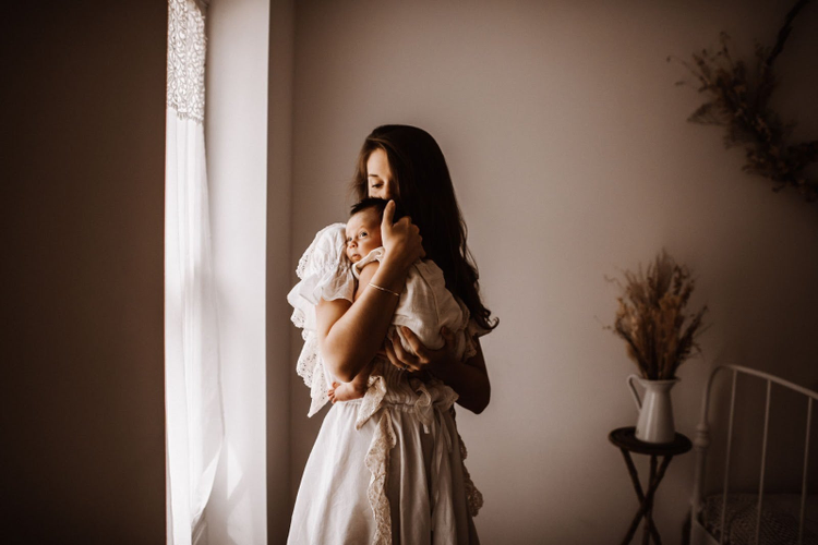 Woman standing in front of window holding a baby. 