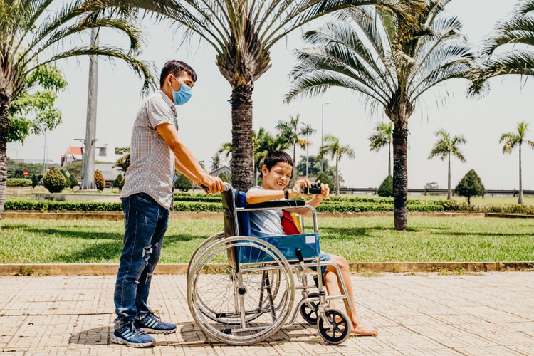 Photo of student pushing another student in a wheelchair while he takes a photo with a camera. 