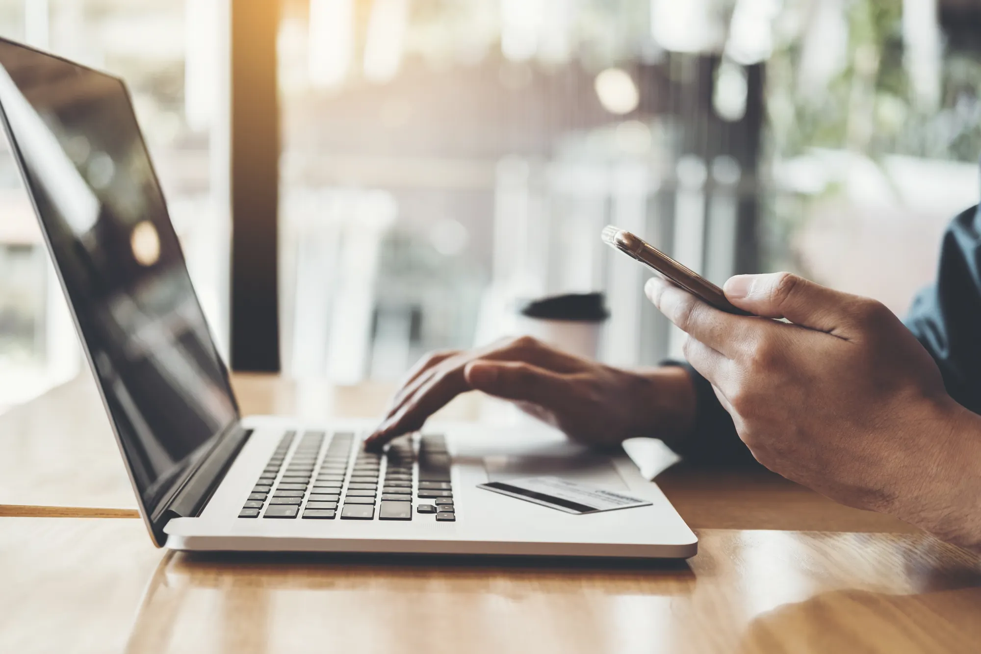 Image of a laptop open with someones hands typing while holding a cell phone in the other hand.