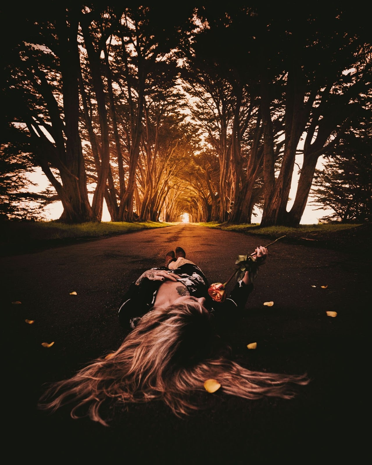 A model smells a rose while lying on a road, illuminated by golden sunrays penetrating the trees in the background.