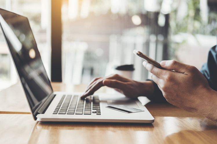 Image of a laptop open with someones hands typing while holding a cell phone in the other hand. 