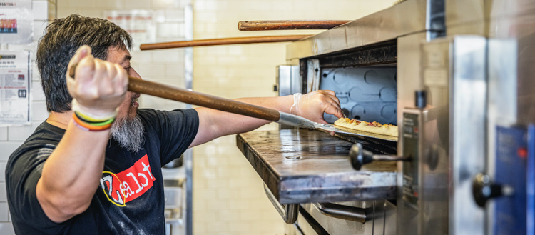 Man putting a pizza in a pizza oven using a wooden paddle. 