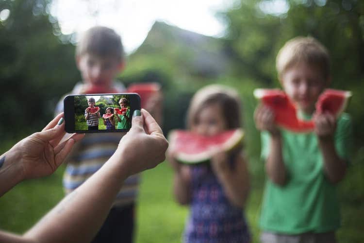 Picture being taken with a cell phone of kids eating watermelon.