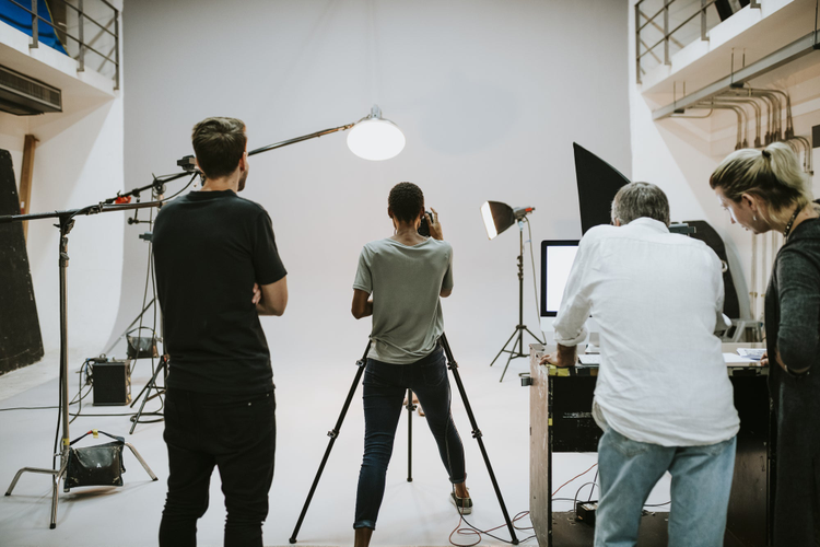 Photography crew working in studio backs facing to the camera amongst cameras and lights looking towards white wall shoot space