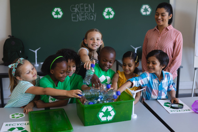 Schoolkids putting bottles in recycle container at desk.