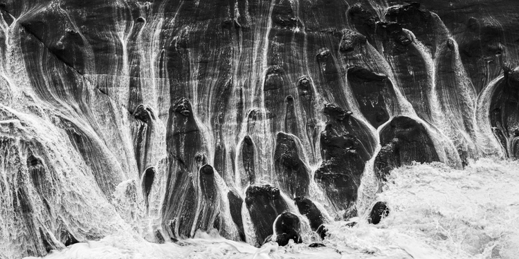 Black and white photo of waves crashing against a large rock at sea
