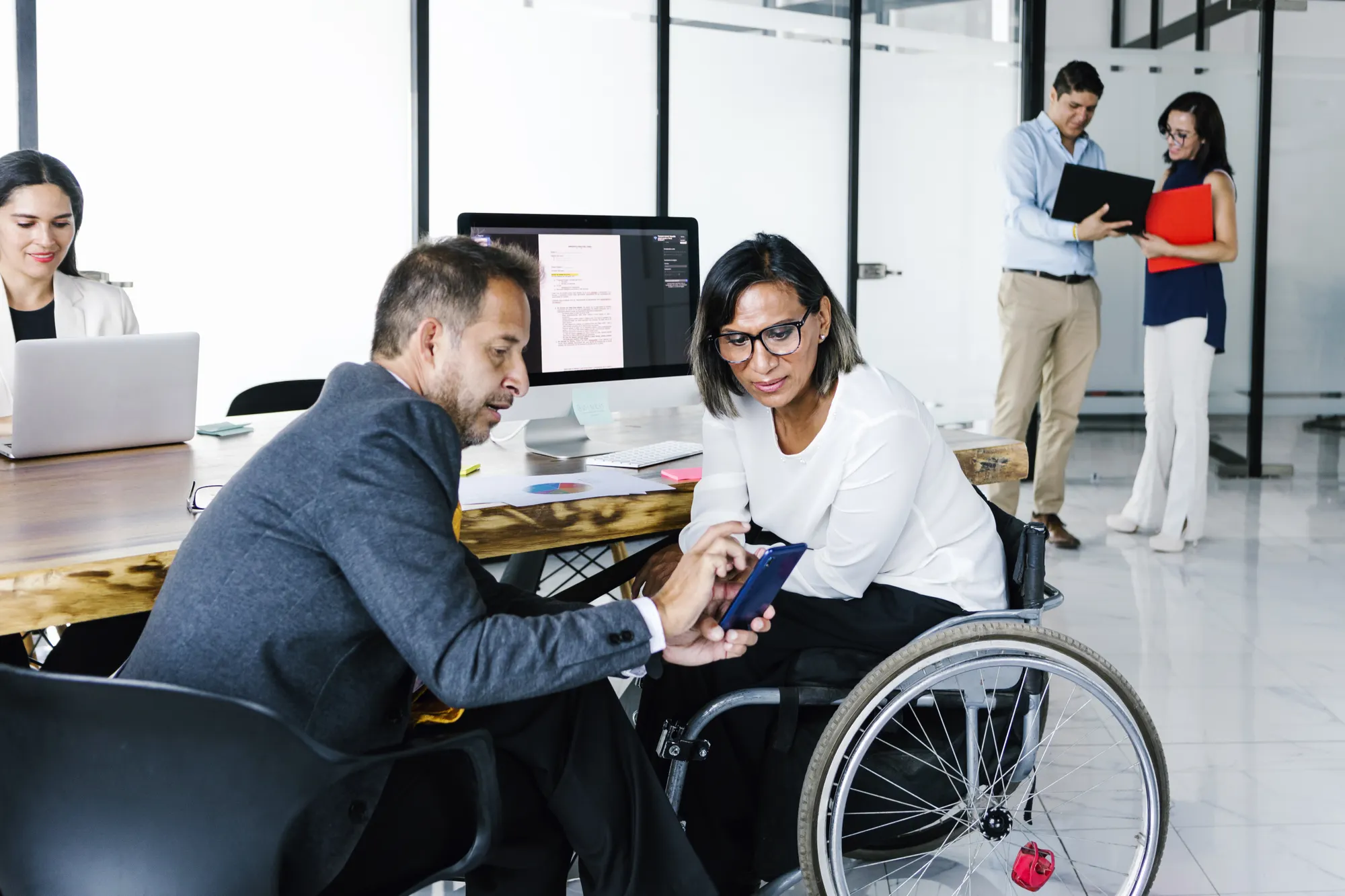 Latinx transgender businesswoman in wheelchair with office colleague checking email on smartphone and office employees working in the background.