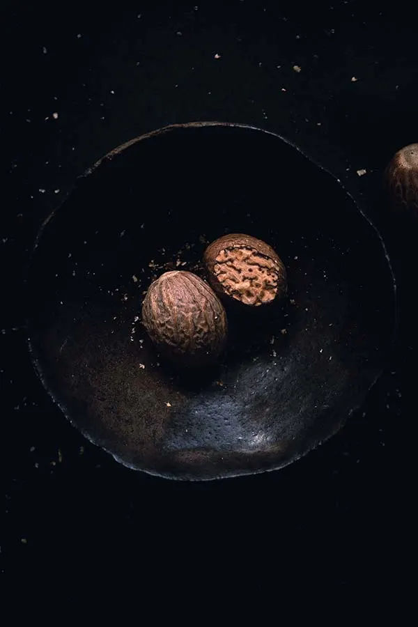 Overhead macro photography of two nutmeg seeds placed inside a ceramic bowl. One seed has been partially grated to expose its interior textures and grain.