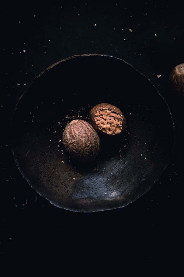 Overhead macro photography of two nutmeg seeds placed inside a ceramic bowl. One seed has been partially grated to expose its interior textures and grain.