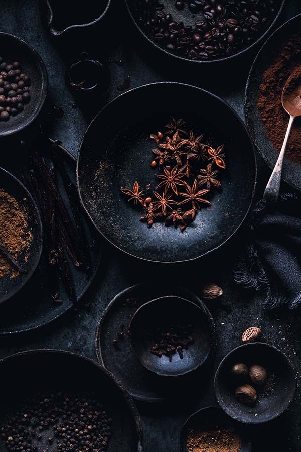 An overhead shot of a collection of different seeds, spices, and beans in earthy, ceramic dishware, atop a black fabric backdrop.