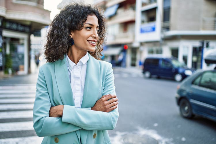 Woman standing on the street next to a crossing, wearing a pale blue suit. She has her arms crossed.