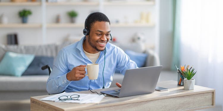 A man in a wheelchair wearing headset, using laptop, drinking coffee at home office.