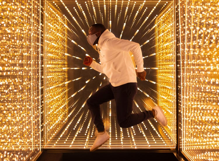 Man wearing mask, jumping between light filled mirrors. 