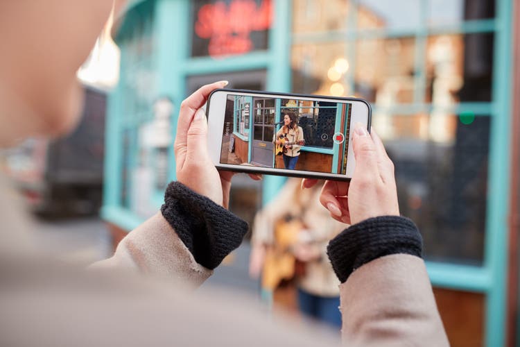 Person Filming Female Musician Busking Playing Acoustic Guitar And Singing To Crowd On Mobile Phone.