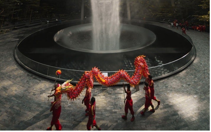Artwork by Pat Kay: A photo of a street square. There is a fountain in the middle. Seven figure in red hold up a Lunar New Year dragon. 