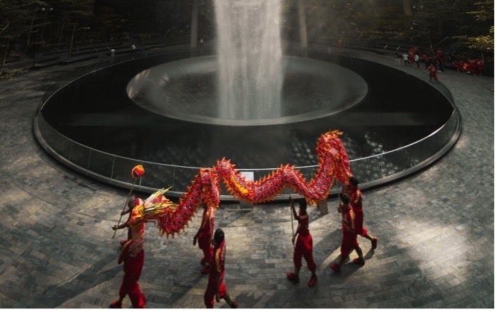 Artwork by Pat Kay: A photo of a street square. There is a fountain in the middle. Seven figure in red hold up a Lunar New Year dragon.