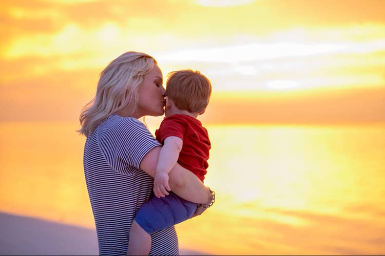 A mother kisses her son's cheek on the beach at sunset.