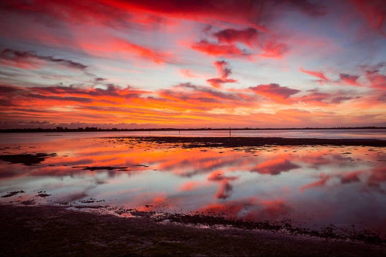 A pre-sunrise photograph at the beach. Pink and orange colors cast the sky, clouds reflected in the water.