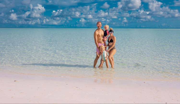 Wide-angle photograph of a family standing in the water at the shoreline of a beach.