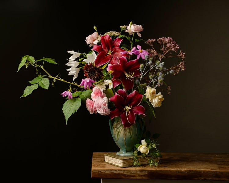 Vase with a variety of flowers perched on a book that is placed on a wooden table. Light falloff produces a natural vignette around the vase.
