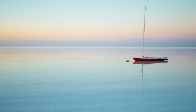 A red and white sailboat anchored in the middle of the ocean. Baby blue sky reflected in the water with a very faint orange glow on the horizon.