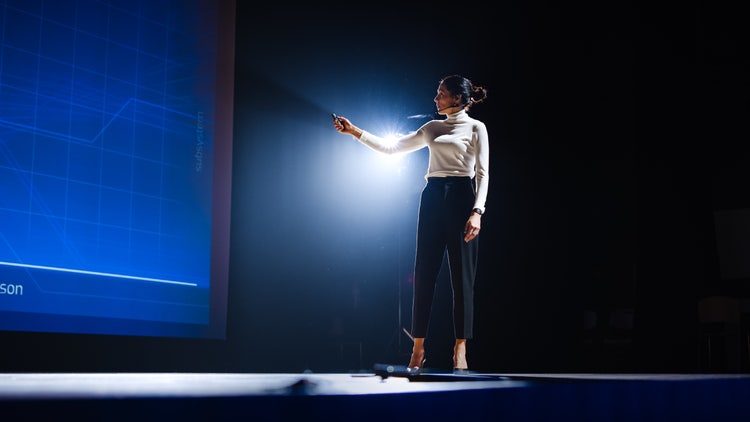 On-Stage Successful Female Speaker Presents Technological Product, Uses Remote Control for Presentation.