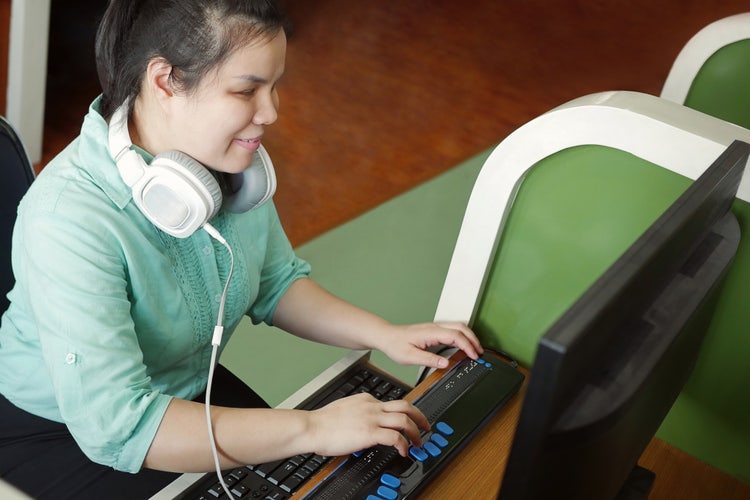 Asian young blind person woman with headphone using computer with refreshable braille display or braille terminal a technology device for persons with visual disabilities.