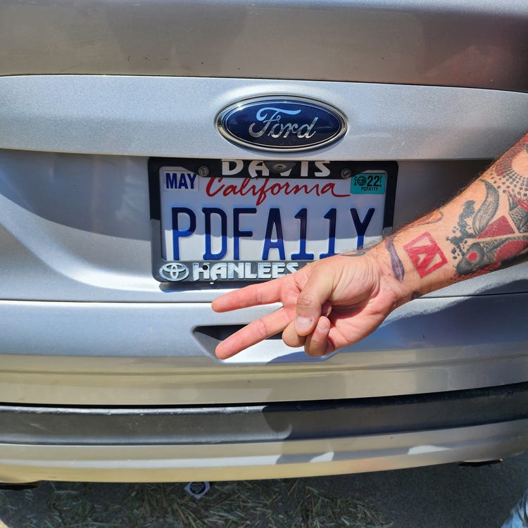 Dax holds out two fingers in a peace sign. His Adobe tattoo is clearly visible amongst his others. His hand is held near his car’s personalized license plate showing the words PDF A11Y.