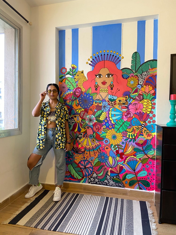 Srishti standing beside a mural of her work, illustrating a woman surrounded by flowers and insects