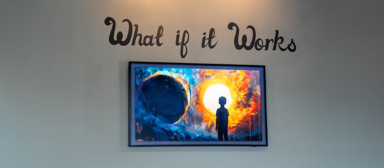 Picture of the words “What if it Works” in cursive over a framed print of a digital, modern-style painting of a person looking at what appears to be two solar bodies. The person is a silhouette on the right in the foreground. On the left in the middle ground is a large planet. On the right in the background is the sun. The sun has a fiery yellow-orange ring, while everything else in the painting is in dark shades of blue.