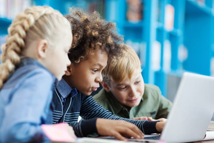 Group of three excited diverse students using laptop sitting at desk in school library.