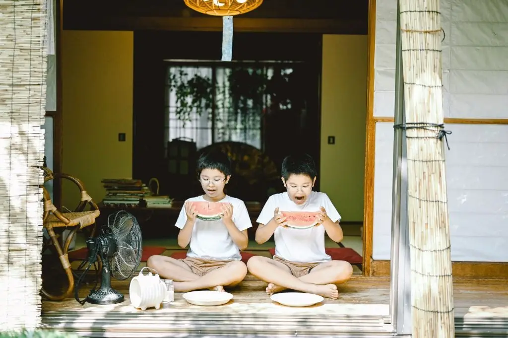 dos niños de tokio sentados frente a un ventilador vestidos de la misma forma comiendose una sandia dividida en la mitad