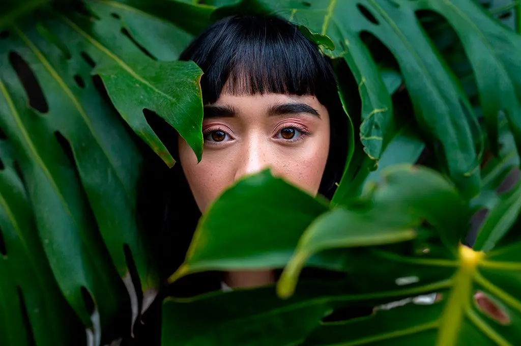  rostro de una joven asiatica detras de unas hojas verdes de un arbol Imagen sin editar