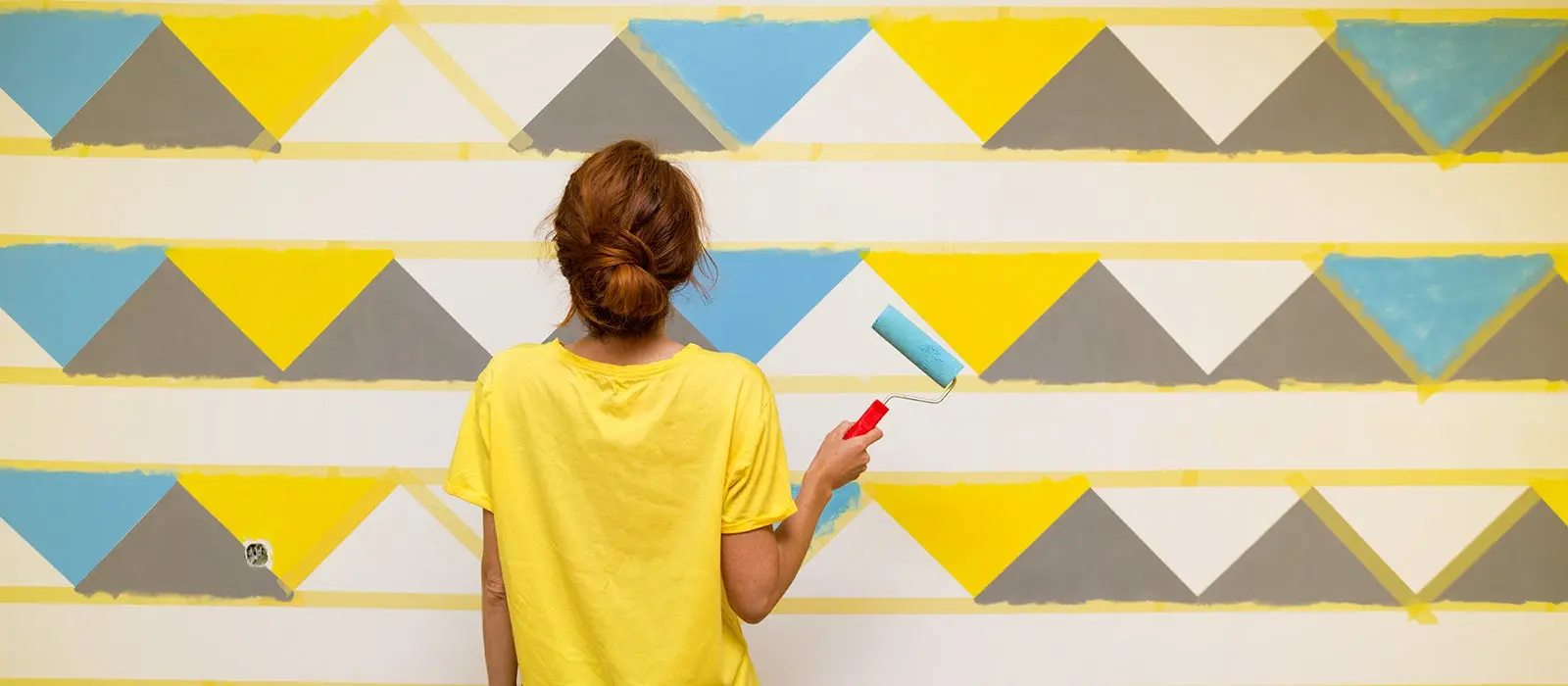 Mujer de espaldas con una camisa amarilla y en su mano derecha un rodillo pintando una pared con formas de triangulos 