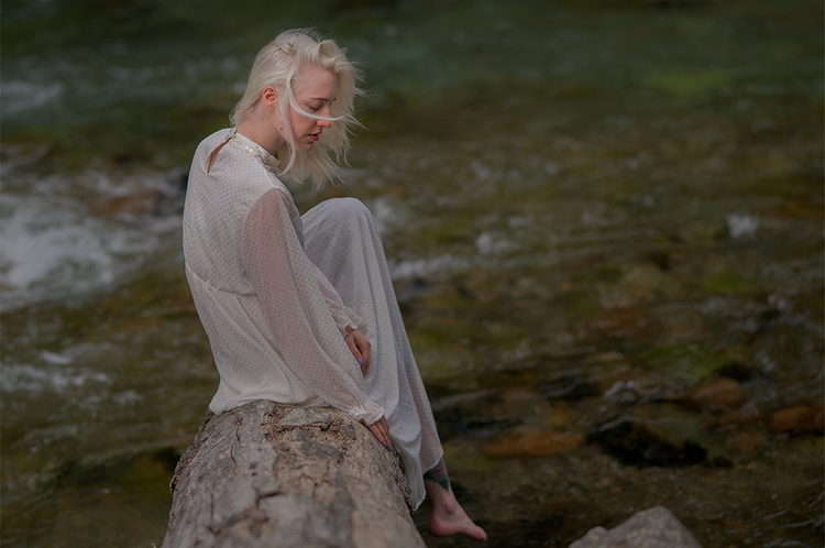 imagen en crudo de una mujer joven de cabellera blanca sentada sobre una roca 