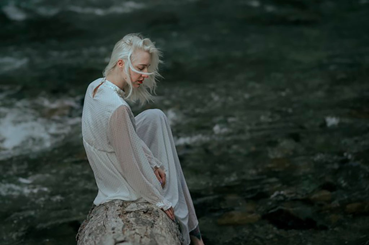 imagen de una joven con colores saturados con prendas y cabellera blanca sentada sobre una roca 