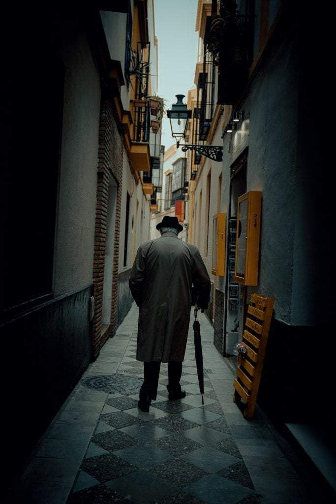 Anciano atravesando un callejon tomando de apoyo una sombrilla, gradiente de color Lightroom