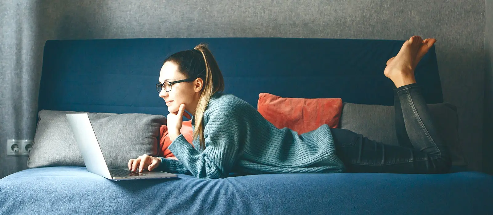 Mujer joven recostada sobre un sofa color azul trabajando en su computador 